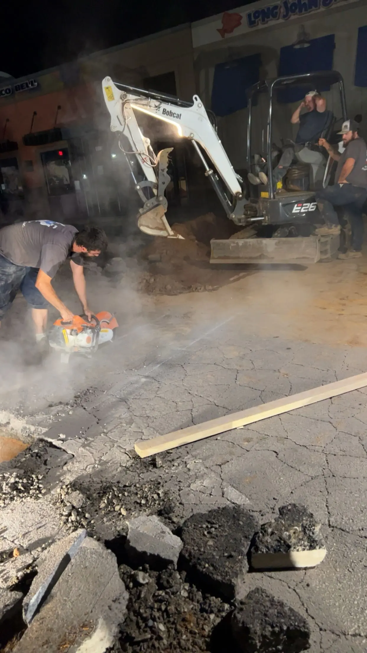 After-Hour Service Nighttime emergency plumbing crew cutting cracked asphalt with a handheld concrete saw while a compact Bobcat excavator digs a pothole in a parking lot, dust and debris rising as workers repair a burst pipe or sewer line.