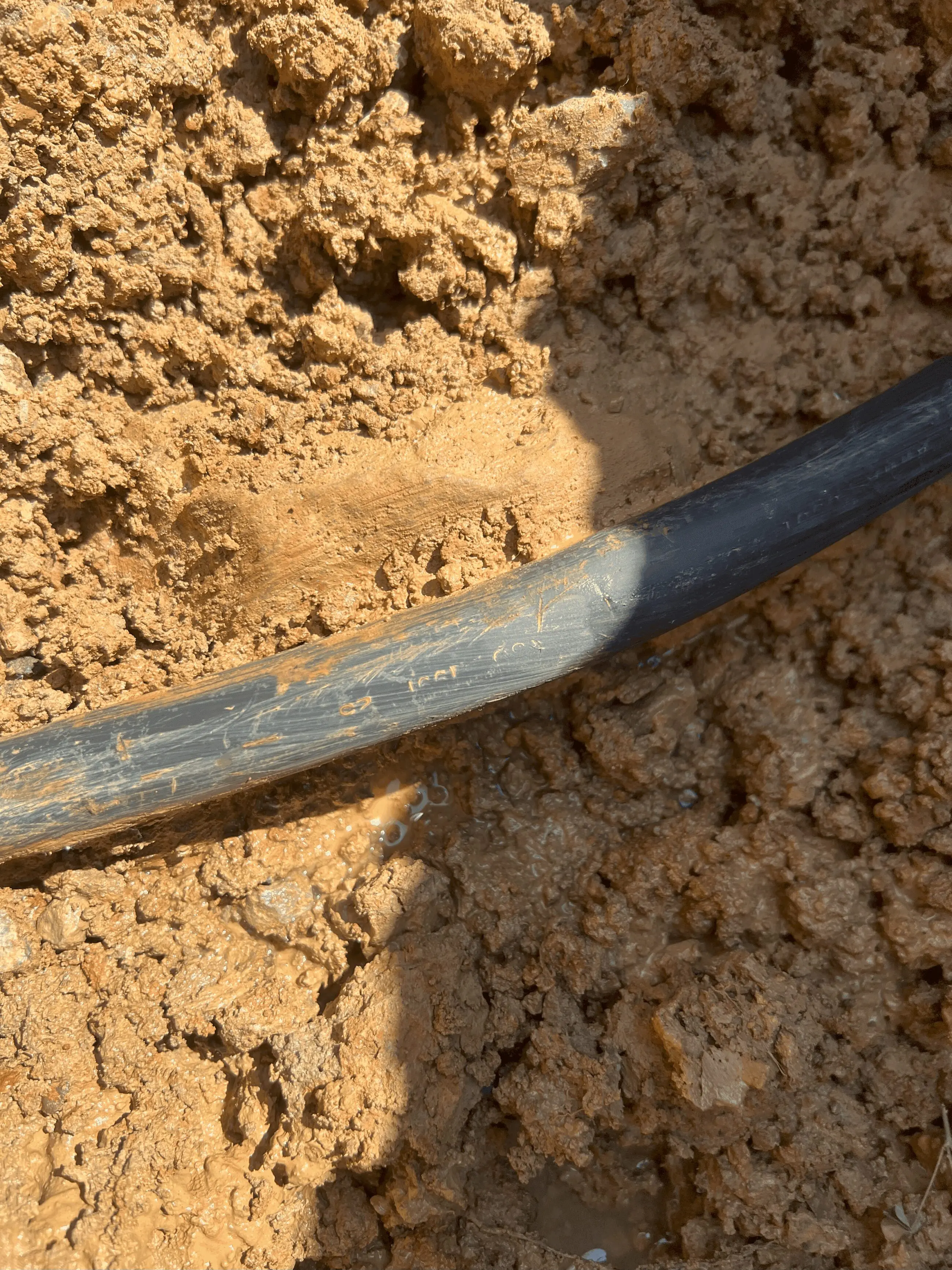 IMG_1978 copy Black polyethylene water service pipe partially exposed in wet brown clay soil, close-up view showing scratches and a small leak where muddy water seeps from the buried plumbing line.