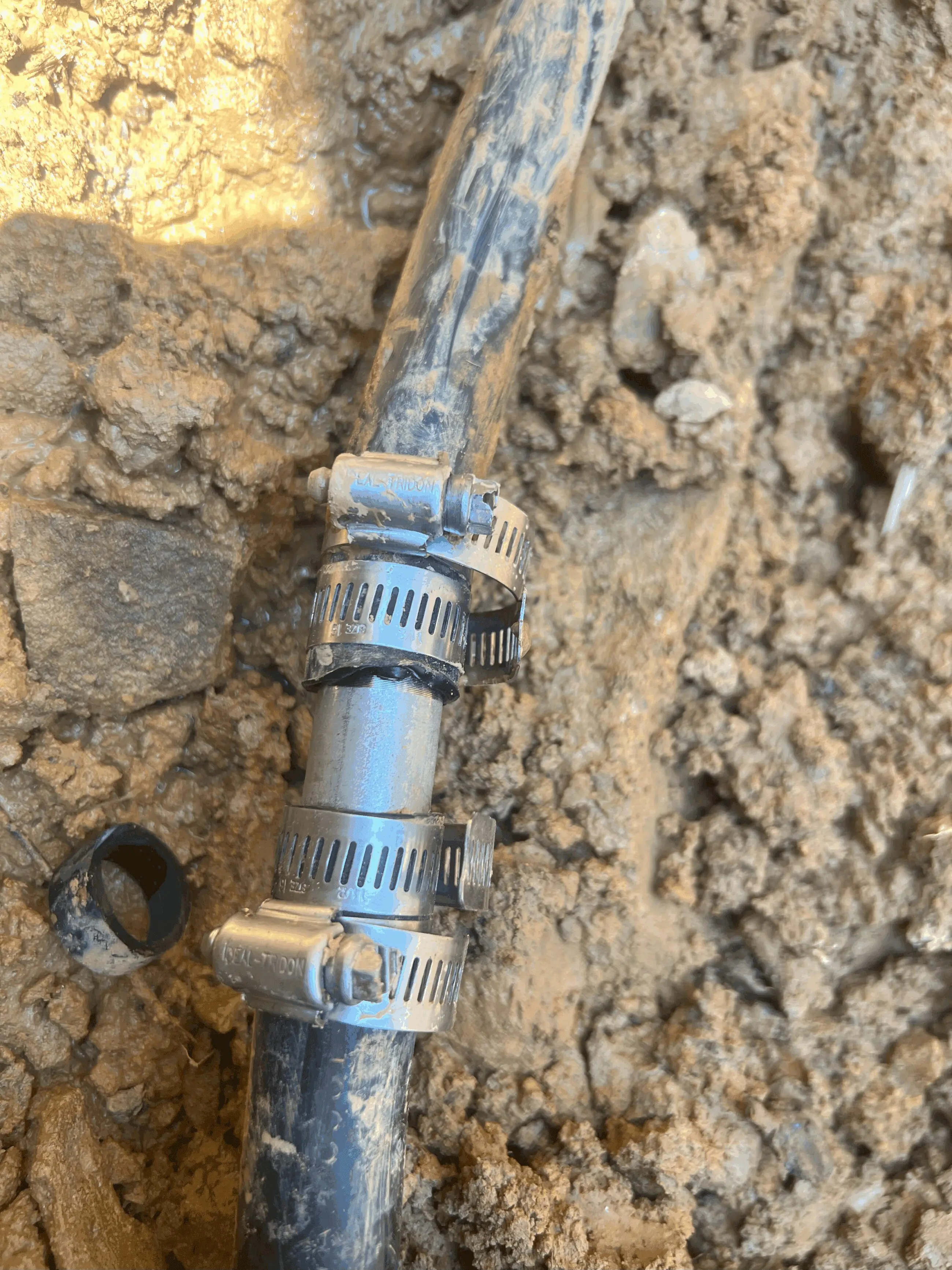 IMG_1980 copy Plastic underground water line in muddy soil with a metal coupling and three stainless steel hose clamps securing a repair, illustrating trench repair and water line replacement services by Walker’s Plumbing in Bedford/Lynchburg/Roanoke.