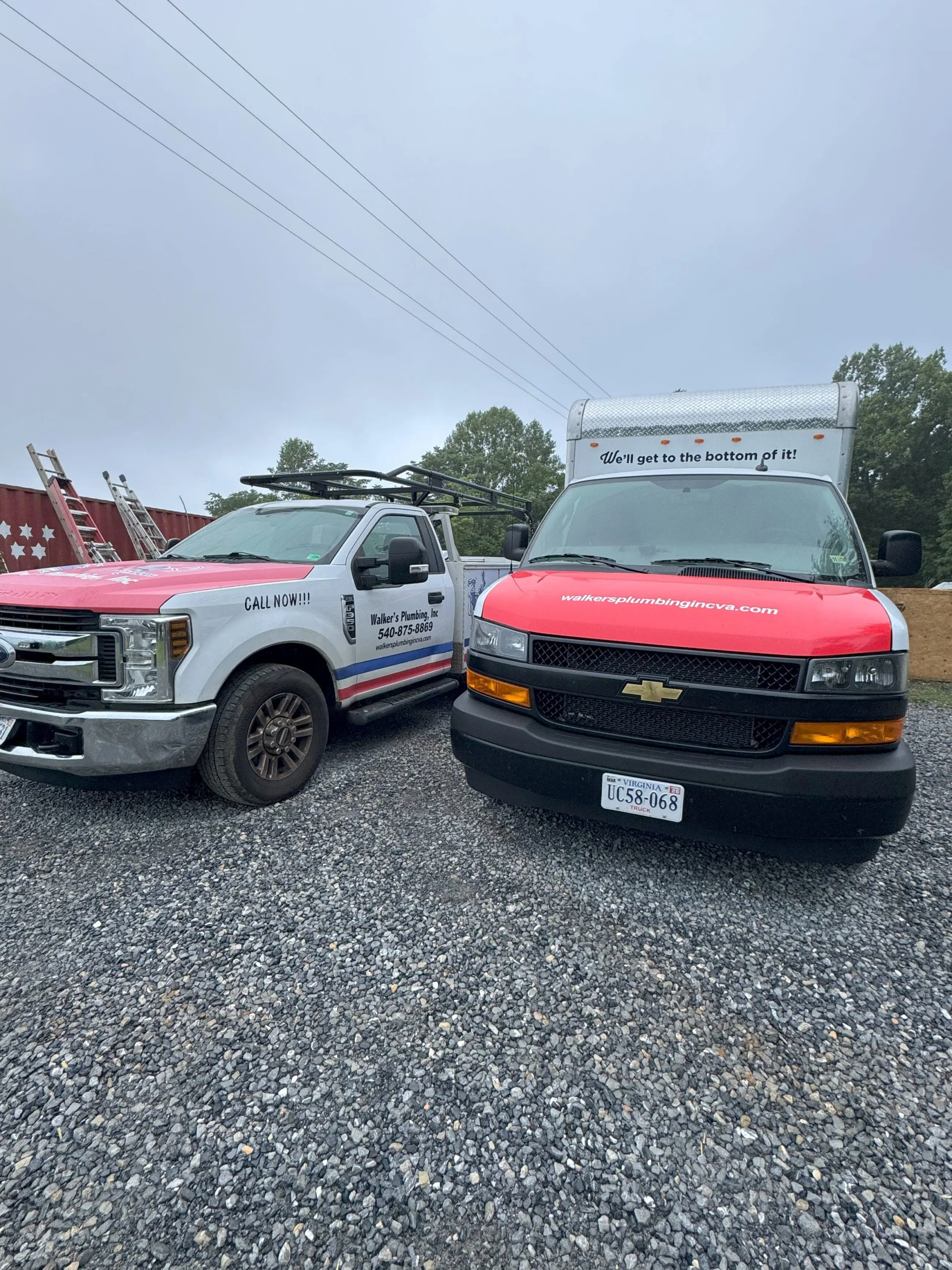 IMG_4444 copy Two Walker’s Plumbing service vehicles parked on gravel — a white pickup with ladder rack and company name and phone on the door next to a red-and-white service van with the company website on the hood and the slogan “We’ll get to the bottom of it!”, under overcast sky.