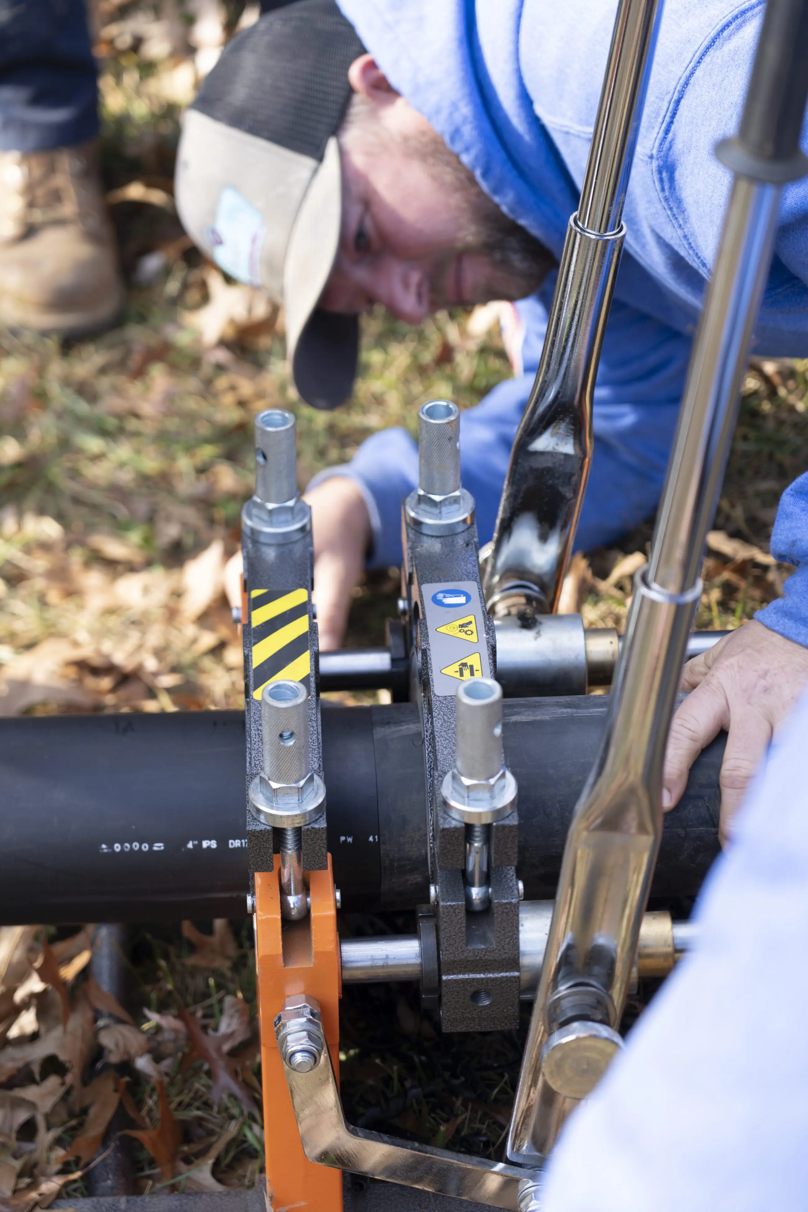 Pipe Bursting Walker-16 Plumber's hands and tools clamping and welding a black plastic water pipe outdoors using a hydraulic fusion machine, showing professional plumbing and water treatment repair work.