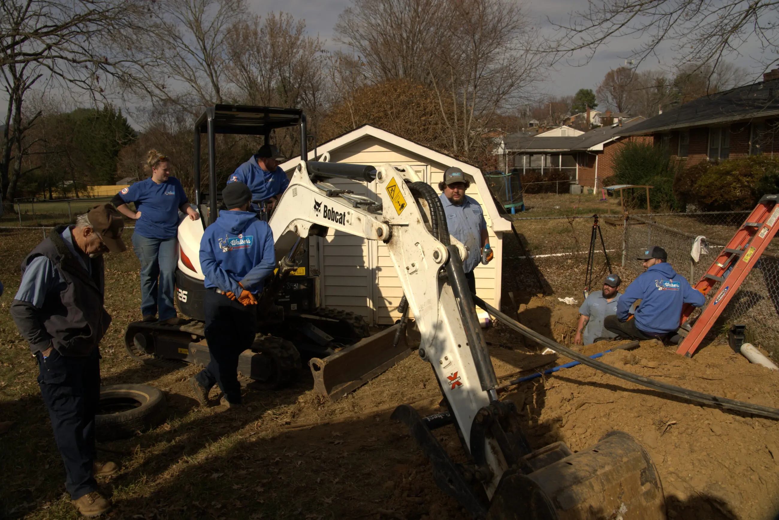 Pipe Bursting Walker-49 Walker’s Plumbing crew in blue uniforms operating a Bobcat mini excavator and working around a backyard trench by a shed, installing or repairing water/well lines with ladders, hoses, and tools visible, illustrating local plumbing and water treatment service.
