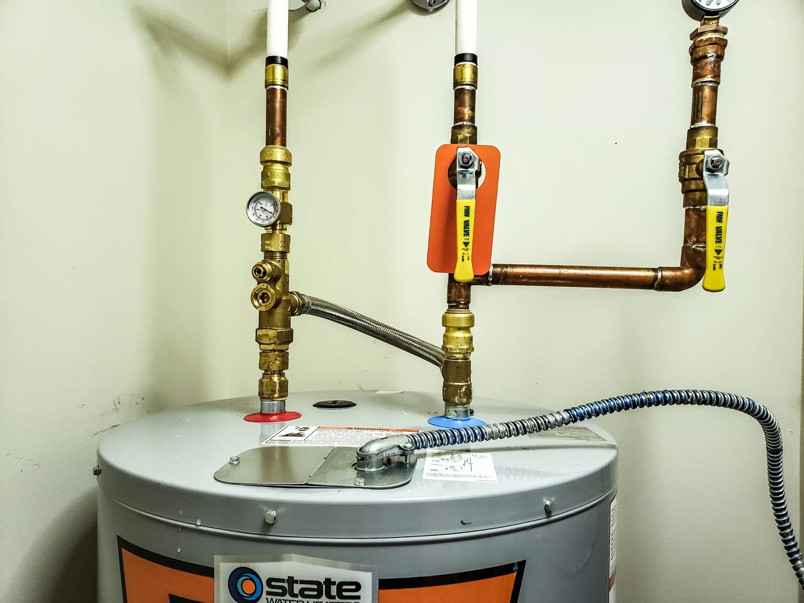 Water Heater Repair Hot water heater tank with copper supply and return pipes, pressure gauge and yellow-handled shutoff valves installed above the unit, flexible stainless water line and electrical conduit — residential water heater service and repair scene for Walker’s Plumbing.