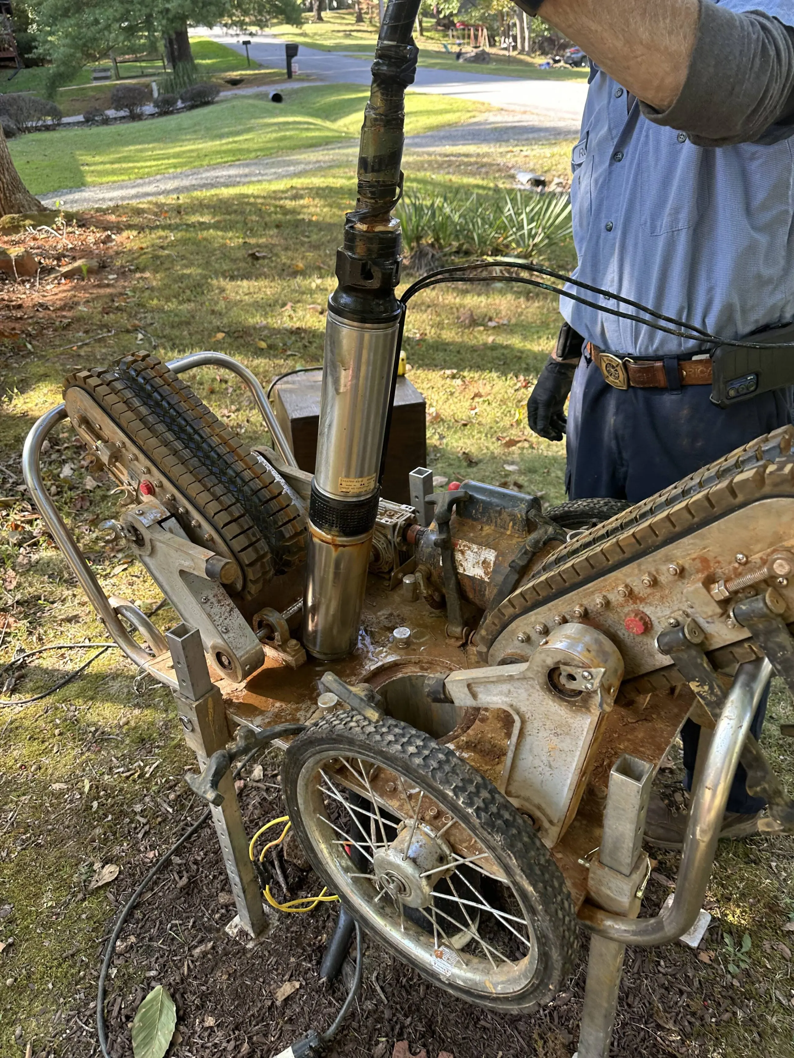 What_Your_Well_Pump_May_Be_Telling_You Technician removing a stainless steel submersible well pump from a portable well-servicing rig on a grassy residential property, showing the pump column, wiring, rig wheels and tools during repair/replacement work for Walker’s Plumbing well pump service.